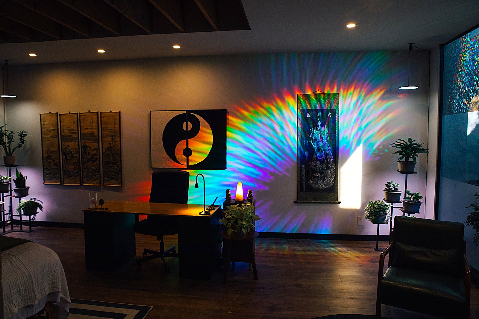 Sunlight through the rainbow window film fills the Gateway Acupuncture treatment room with color, beside the yin-yang painting and a lit candle